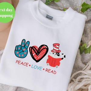 May include: White sweatshirt with a graphic design featuring a peace sign, a red heart, and a cat reading a book. The text reads "PEACE LOVE READ" in red and blue. The sweatshirt is a Gildan brand.