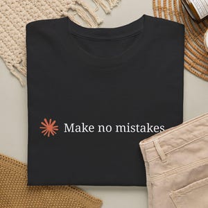 May include: A black short-sleeved t-shirt featuring a small coral-colored sunburst icon on the left side and the white text "Make no mistakes" printed in a clean, sans-serif font. The shirt is folded neatly, showcasing the graphic design clearly.