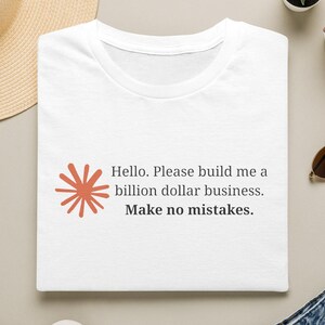May include: A white t-shirt featuring a rust-colored starburst graphic and black text that reads: Hello. Please build me a billion dollar business. Make no mistakes. The shirt is displayed alongside a straw hat, sunglasses, and blue jeans.