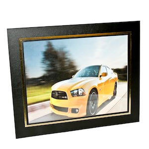 May include: A framed print featuring a vibrant yellow sports car speeding down a road. The image is set within a black frame with a gold inner border, creating a striking contrast. The car's motion is emphasized by the blurred background.