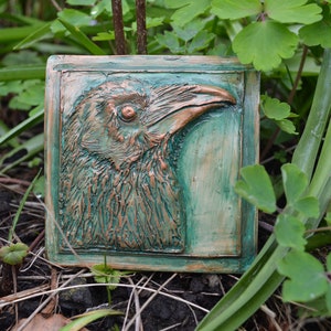 May include: A close-up of a ceramic tile featuring a raised relief sculpture of a raven's head. The raven is facing right and has a copper-coloured finish. The tile is set against a green background.