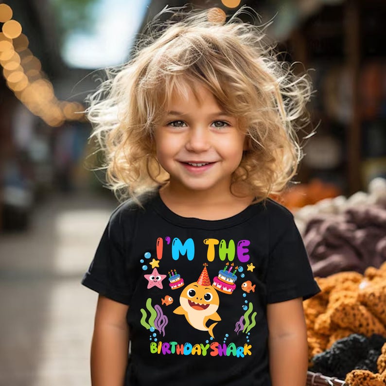 Puede incluir: Una camiseta negra de manga corta con un gr&aacute;fico colorido de un tibur&oacute;n de dibujos animados con un gorro de fiesta, rodeado de dos pasteles de cumplea&ntilde;os, una estrella de mar y algas. El texto dice I'M THE BIRTHDAY SHARK en letras brillantes.
