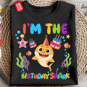 Puede incluir: Una camiseta negra con un gr&aacute;fico colorido de un tibur&oacute;n de dibujos animados con un gorro de fiesta, rodeado de dos pasteles de cumplea&ntilde;os, una estrella de mar, dos peces naranjas peque&ntilde;os y algas verdes y moradas. El texto dice I'M THE BIRTHDAY SHARK.