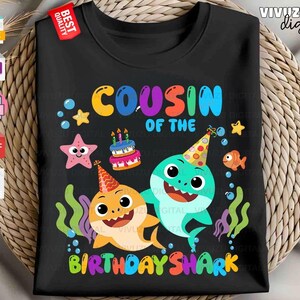 Puede incluir: Una camiseta negra con un gr&aacute;fico colorido y el texto "COUSIN OF THE BIRTHDAY SHARK". El dise&ntilde;o incluye dos tiburones de dibujos animados con gorros de fiesta, un pastel de cumplea&ntilde;os, una estrella de mar, un pez naranja, burbujas y algas.