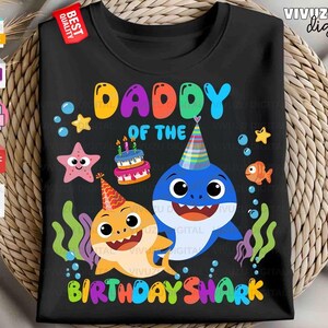 Puede incluir: Una camiseta negra con un gr&aacute;fico colorido de dos tiburones con gorros de fiesta, un pastel de cumplea&ntilde;os, una estrella de mar y un pez. El texto DADDY OF THE BIRTHDAY SHARK aparece en letras multicolores, rodeado de burbujas y algas.