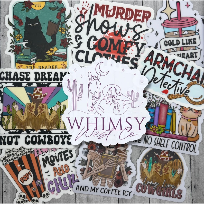 May include: A collection of colorful stickers with various designs. One sticker features a black cat reading a book, another says "Murder Shows & Comfy Clothes." Other stickers include a cowboy, a skeleton, and the text "Whimsy West Co."