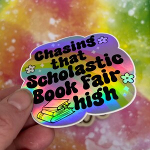 May include: A holographic sticker in the shape of a cloud with the text "Chasing that Scholastic Book Fair high" in black. The sticker has a rainbow design and small flower and diamond accents.