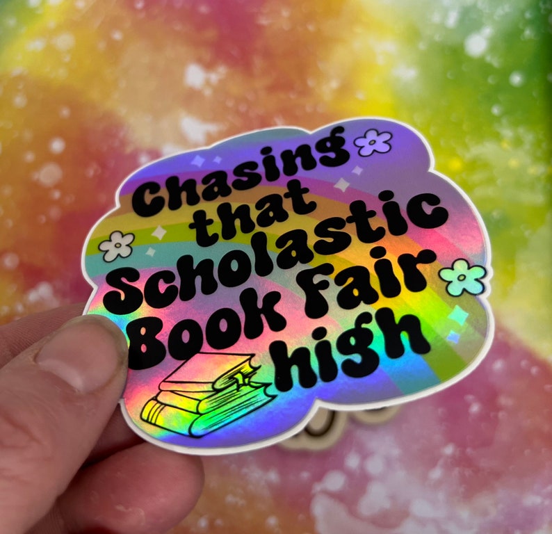 May include: A holographic sticker in the shape of a cloud with the text "Chasing that Scholastic Book Fair high" in black. The sticker has a rainbow design and small flower and diamond accents.