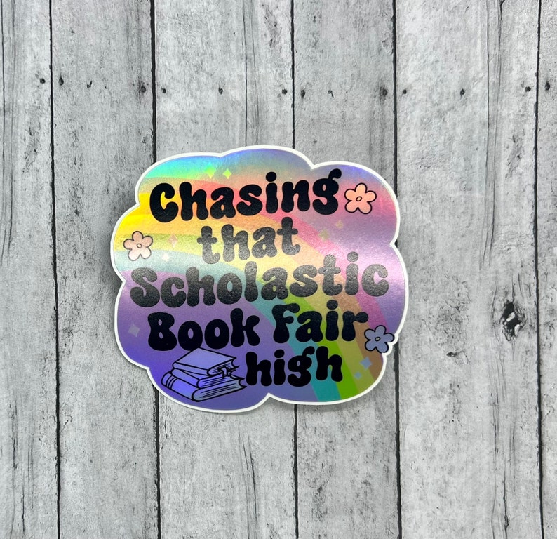 May include: A holographic sticker with the text "Chasing that Scholastic Book Fair high" in black, with a cloud-like shape and flower accents. The sticker features a stack of books at the bottom.