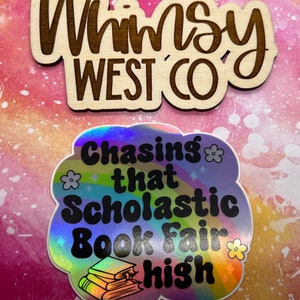 May include: Two stickers on a pink and white background. The top sticker is wooden with the text "Whimsy West Co" in a dark brown color. The bottom sticker is holographic with the text "Chasing that Scholastic Book Fair high" and a stack of books.