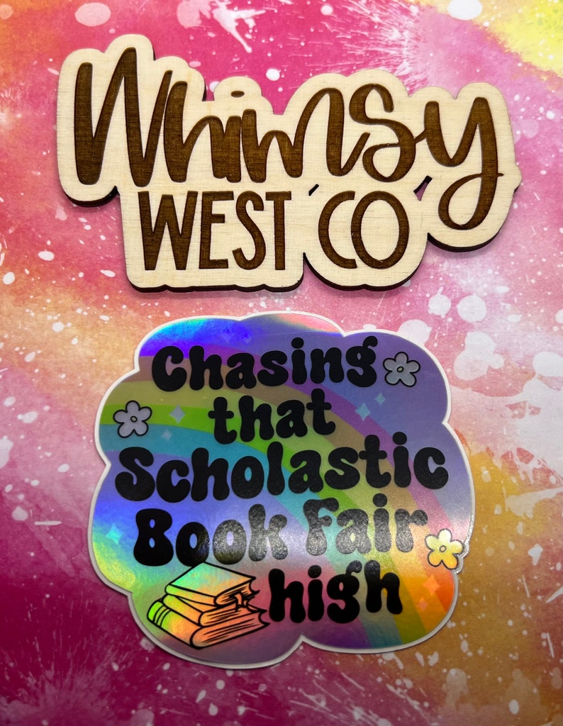 May include: Two stickers on a pink and white background. The top sticker is wooden with the text "Whimsy West Co" in a dark brown color. The bottom sticker is holographic with the text "Chasing that Scholastic Book Fair high" and a stack of books.