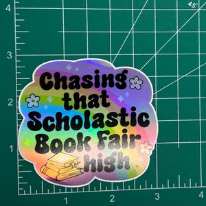 May include: A holographic sticker with the text "Chasing that Scholastic Book Fair high." The sticker has a cloud shape with a rainbow design, flowers, and diamond shapes. A stack of books is at the bottom.