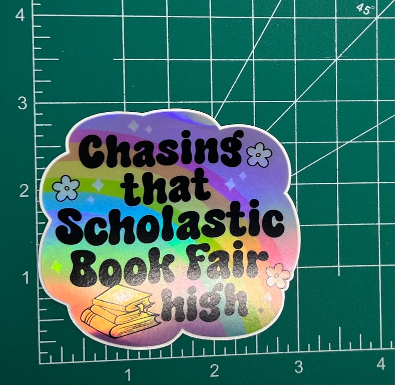 May include: A holographic sticker with the text "Chasing that Scholastic Book Fair high." The sticker has a cloud shape with a rainbow design, flowers, and diamond shapes. A stack of books is at the bottom.