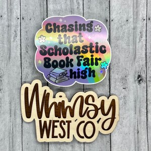 May include: Two stickers on a wooden surface. The top sticker is cloud-shaped with the text "Chasing that Scholastic Book Fair high" and a stack of books. The bottom sticker says "Whimsy West Co."