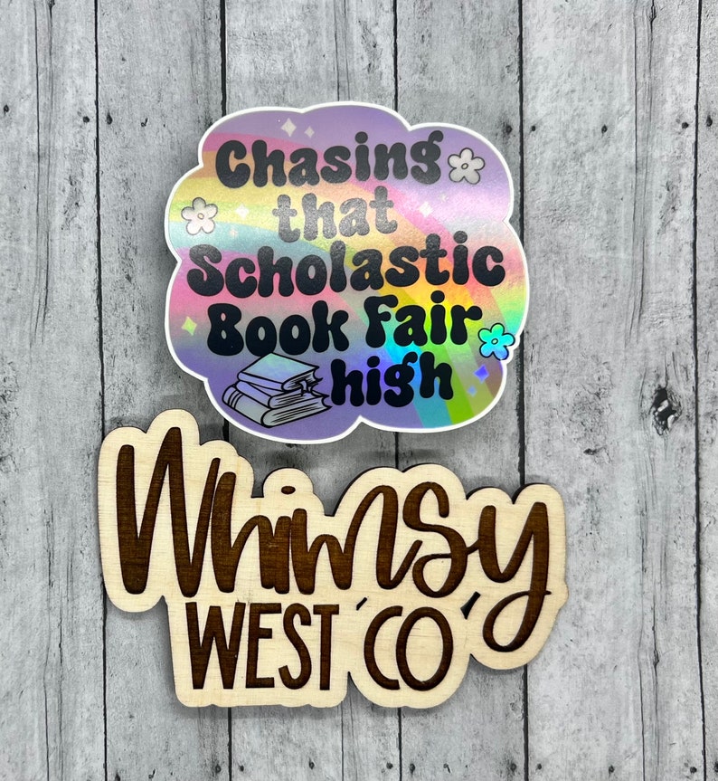 May include: Two stickers on a wooden surface. The top sticker is cloud-shaped with the text "Chasing that Scholastic Book Fair high" and a stack of books. The bottom sticker says "Whimsy West Co."