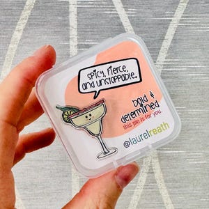 Op de afbeelding: Een transparante plastic doos met een roze en witte emaille pin erin. De pin heeft een cartoon margarita glas met een limoenpartje en een glimlachend gezicht. De tekst op de doos luidt "Spicy, Fierce, and Unstoppable. Bold & Determined this pin is for you. @laurelreath"