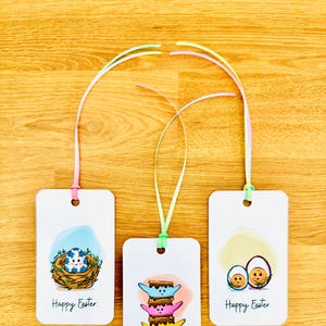 Könnte beinhalten: Drei Oster-Geschenk-Etiketten mit bunten Bändern. Die Etiketten zeigen Illustrationen eines Nestes mit einem Ei, einem Stapel Marshmallow-S'mores und zwei Eiern in einem Nest. Jedes Etikett trägt den Text "Frohe Ostern."