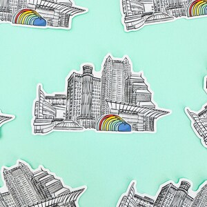 Puede incluir: Un conjunto de cinco pegatinas que muestran una ilustraci&oacute;n en blanco y negro de un horizonte de ciudad con un arco&iacute;ris sobre los edificios. Las pegatinas est&aacute;n sobre un fondo azul claro.