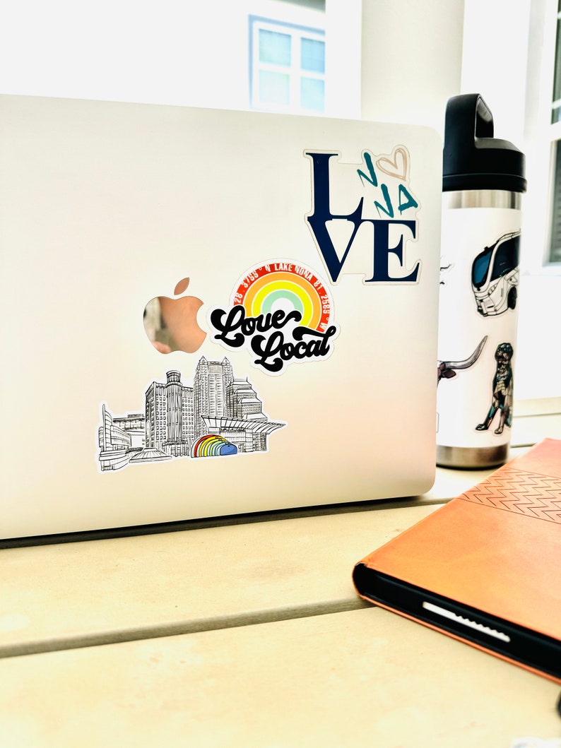 Puede incluir: Un port&aacute;til plateado con varias pegatinas en la tapa. Las pegatinas incluyen un arco&iacute;ris con el texto "Love Local", un horizonte de ciudad y una pegatina azul "LOVE". Un diario de cuero marr&oacute;n est&aacute; sobre la mesa junto al port&aacute;til.