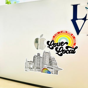 Puede incluir: Una ilustraci&oacute;n en blanco y negro de un horizonte de ciudad con un arco&iacute;ris y el texto "Love Local" en una fuente retro. El texto "28.5765&deg; N LAKE NONA FL 32833" tambi&eacute;n est&aacute; incluido en el dise&ntilde;o.