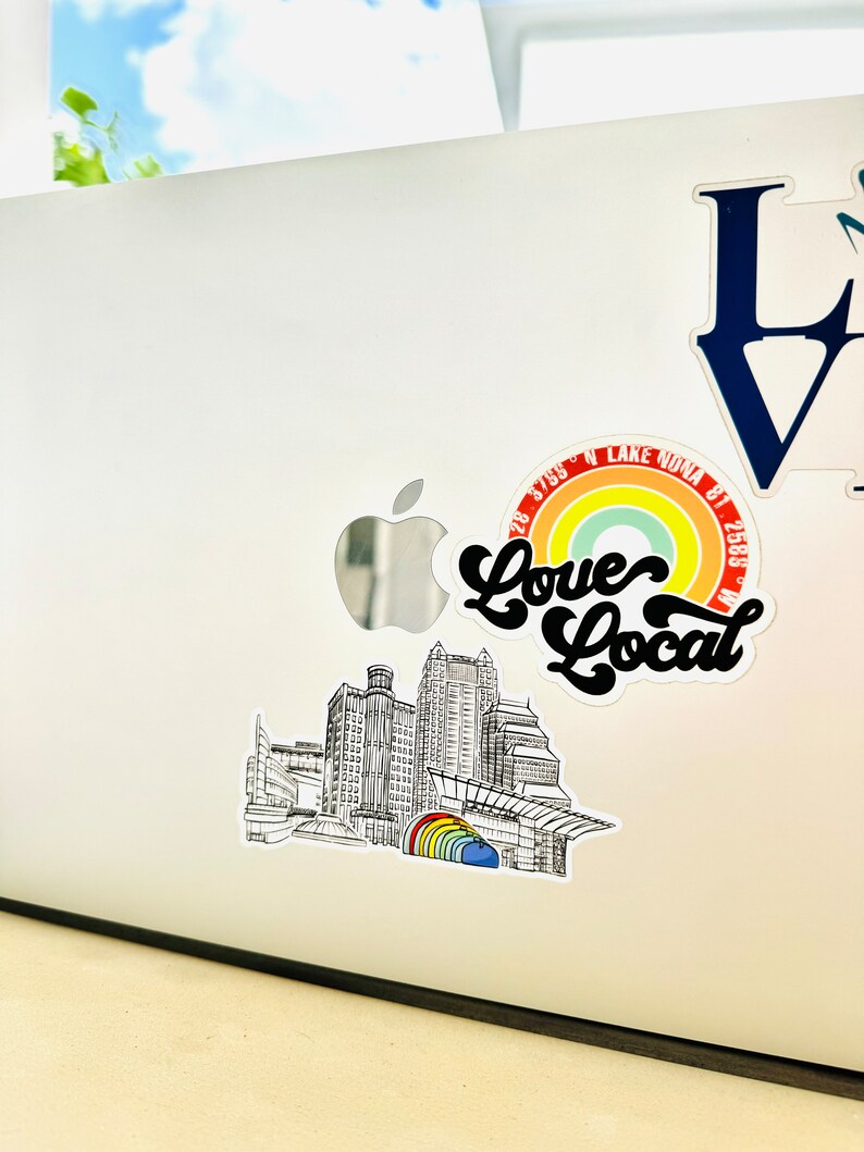 Puede incluir: Una ilustraci&oacute;n en blanco y negro de un horizonte de ciudad con un arco&iacute;ris y el texto "Love Local" en una fuente retro. El texto "28.5765&deg; N LAKE NONA FL 32833" tambi&eacute;n est&aacute; incluido en el dise&ntilde;o.