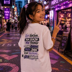 May include: A white t-shirt featuring a graphic print on the back with the names Kim Namjoon, Kim Seokjin, Min Yoongi, Jung Hoseok, Park Jimin, Kim Taehyung, and Jeon Jungkook in purple and black stylized text, accented with small hearts and stars.