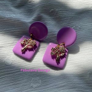 以下が含まれることがあります： 正方形のベースと円形のトップを備えた、ライラック色のイヤリングのペア。各イヤリングは、クリアクリスタルのアクセントが付いた金色の花の模様が特徴です。イヤリングは白いテクスチャのある表面に表示されています。「Fabulava Design」というテキストが表示されています。