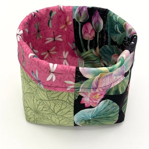 May include: A fabric storage basket with a patchwork design. The interior and top trim feature a pink fabric with white dragonflies. The exterior has green lotus leaf and black floral patterns. The basket is open and empty.