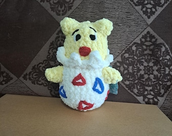 May include: A handmade yellow crochet Togepi plush toy with a white body featuring red and blue triangle accents. The plush has a red nose, black eyes, and a white, fluffy collar. The toy is sitting on a brown surface.