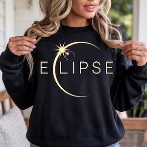 Pode incluir: Camisola preta com a palavra "ECLIPSE" em letras douradas, acompanhada por um sol e uma lua crescente dourados. O design está centrado na frente da camisola.