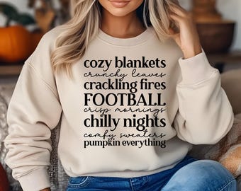 May include: A beige sweatshirt with black text that reads "cozy blankets, crunchy leaves, crackling fires, FOOTBALL, crisp mornings, chilly nights, comfy sweaters, pumpkin everything".
