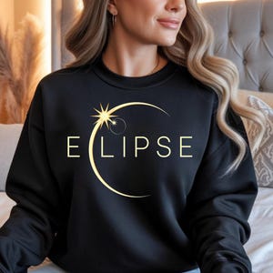 May include: Black sweatshirt with the word "E L I P S E" in a light gold colour. The design features a stylized sun and crescent moon graphic. The sweatshirt is a crew neck style.