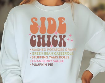 Può includere: Una felpa blu chiaro con il testo "SIDE CHICK" in un font retrò. Il testo è in arancione e marrone con stelle rosa. Sotto il testo c'è un elenco di contorni: purè di patate con sugo, casseruola di fagiolini, involtini di patate dolci ripieni, salsa di mirtilli rossi e torta di zucca.