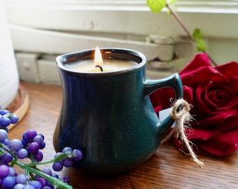 May include: A teal blue ceramic pitcher with a candle burning inside. The candle is made of a light brown wax and has a white wick. The pitcher has a handle and is sitting on a wooden surface.