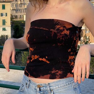 Può includere: Un top a tubo nero e arancione tie-dye con chiusura a bottoni laterale. Il top è realizzato in tessuto a coste e abbinato a jeans di denim chiaro. Il top ha un design unico e artistico.