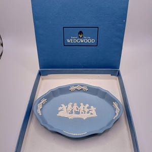 Puede incluir: Un plato de alhajas de jaspe azul Wedgwood con un camafeo blanco del diseño 'Dancing Hours'. El plato está en una caja azul con el logotipo de Wedgwood.