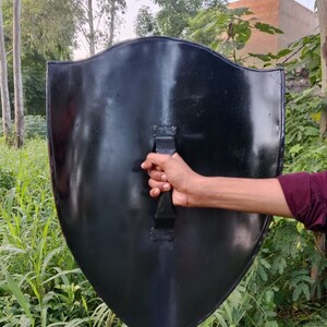 May include: A black metal shield with a handle. The shield is held by a hand in front of a green grassy background.