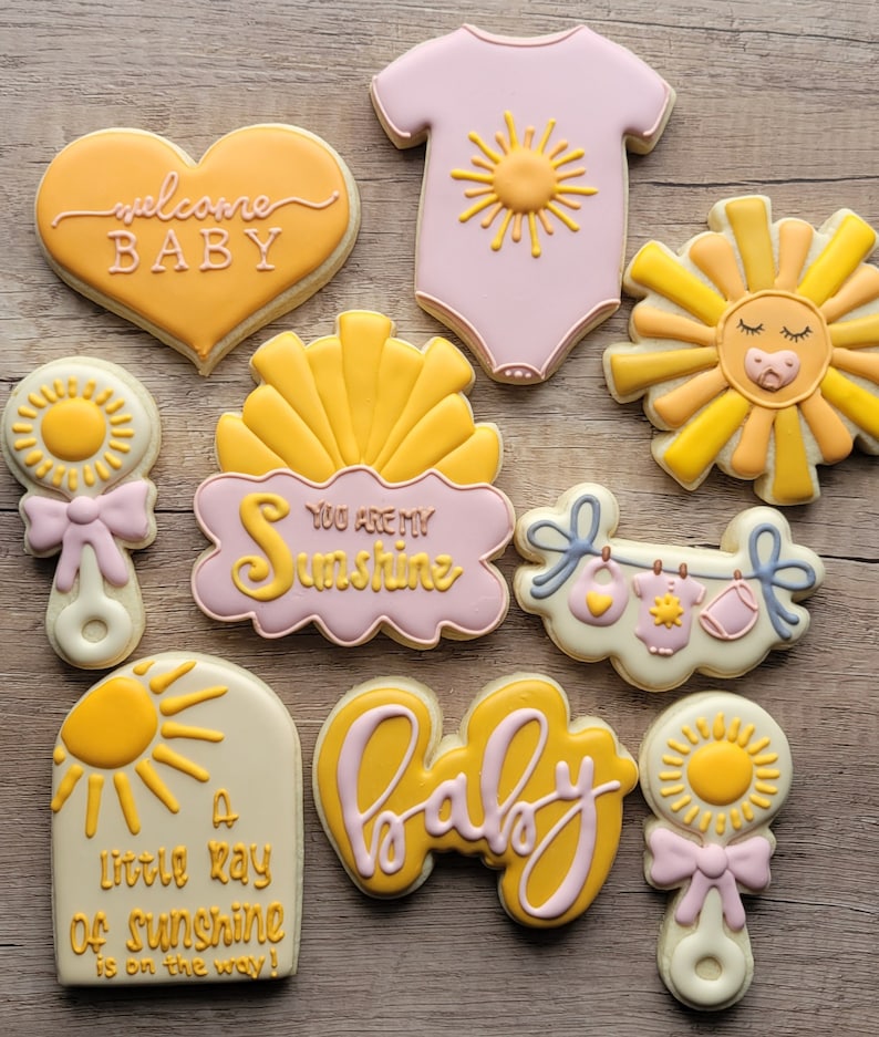 May include: A set of nine decorated sugar cookies with a baby shower theme. The cookies are decorated with yellow, pink, and white icing and feature various baby-related designs, including a sun, a rattle, a onesie, and the words "welcome baby" and "you are my sunshine".