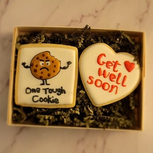 May include: A gift box containing two decorated sugar cookies. One cookie is shaped like a heart and says "Get well soon" in red icing. The other cookie is round and features a cartoon cookie with a grumpy expression and the text "One Tough Cookie".
