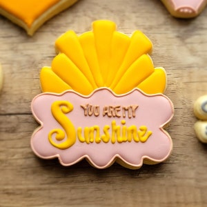 May include: A pink and yellow cookie decorated with a sun and the text "You are my sunshine".