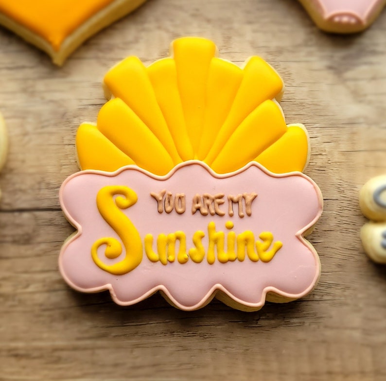 May include: A pink and yellow cookie decorated with a sun and the text "You are my sunshine".