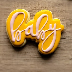 May include: A yellow cookie decorated with pink icing in the shape of the word "baby".