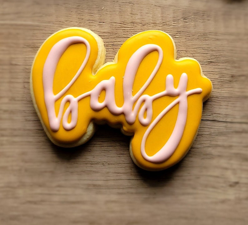 May include: A yellow cookie decorated with pink icing in the shape of the word "baby".