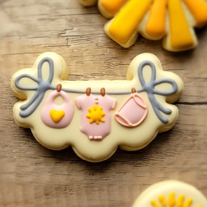 May include: A yellow and white cookie decorated with a clothesline and three baby items: a bib, a onesie, and a diaper. The items are pink and yellow with a sun design.