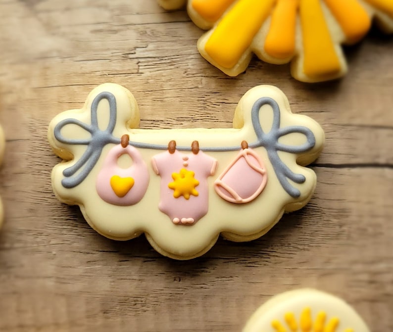 May include: A yellow and white cookie decorated with a clothesline and three baby items: a bib, a onesie, and a diaper. The items are pink and yellow with a sun design.