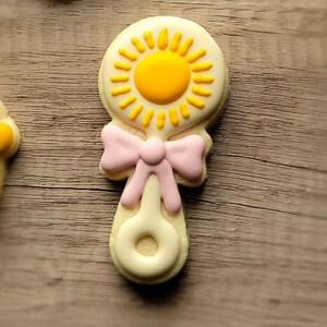 May include: A yellow and pink baby rattle cookie decorated with a sun design.