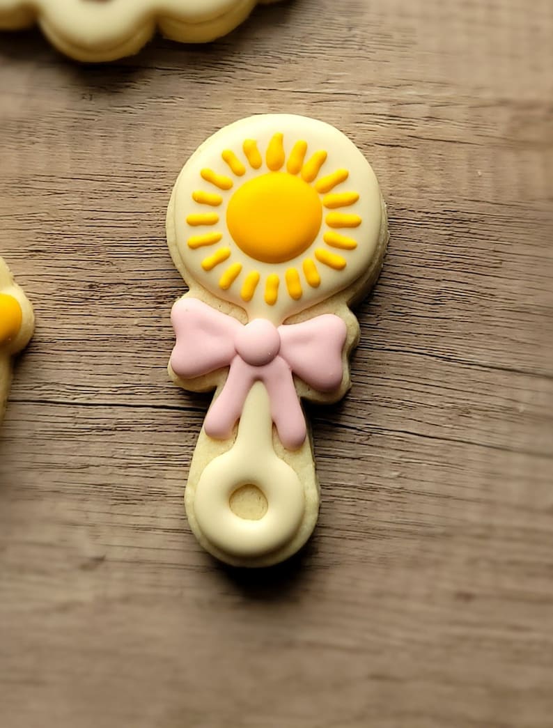 May include: A yellow and pink baby rattle cookie decorated with a sun design.