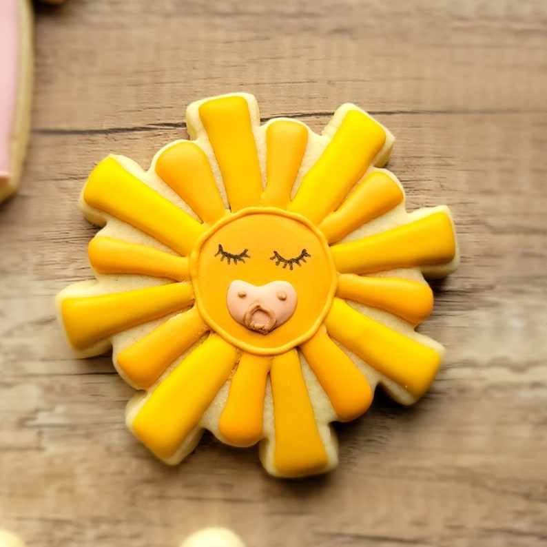 May include: A yellow sugar cookie decorated with a sun face with closed eyes and a pacifier. The cookie is shaped like a sun with rays.