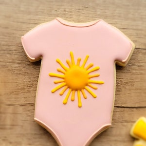 May include: Pink sugar cookie decorated with a yellow sun design, shaped like a baby onesie.
