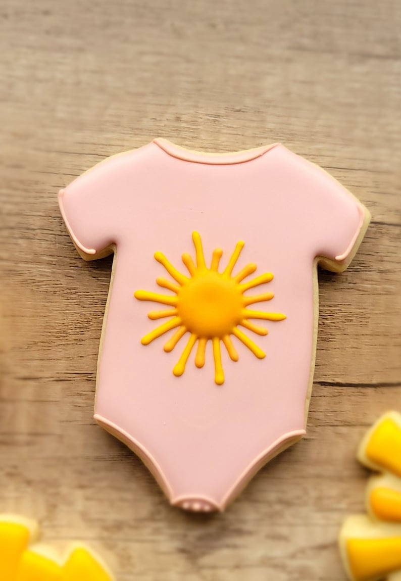 May include: Pink sugar cookie decorated with a yellow sun design, shaped like a baby onesie.